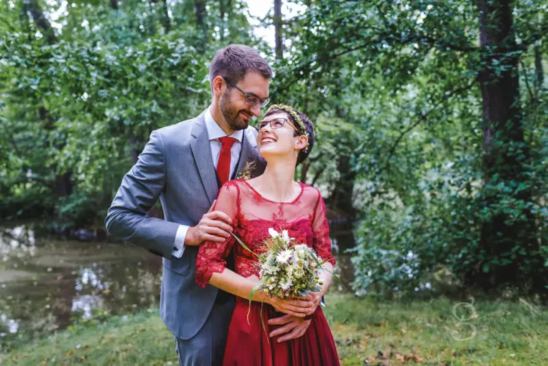 Individuelle Familienshootings & Hochzeitsfotografie vom Profi 9 Brautpaar in einem Park stehend. Die Braut in einem roten, bauchfreien Spitzenkleid steht mit dem Rücken vor ihrem Bräutigam und schaut zu ihm nach oben. Sie hält ihren Brautstrauß in beiden Händen.